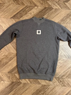 Grå RAW sweatshirt  - Mysig grå sweatshirt med rund halsringning och ribbade muddar vid ärmslut och nederkant. På bröstet sitter en vit patch med svart logga. Perfekt till ett avslappnat streetwear-look.