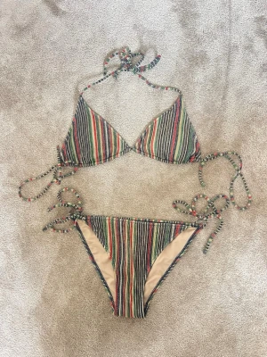 Randig bikini med glitter i flera färger - Säljer en snygg trekantsbikini med färgglada ränder i glittrigt grönt, rött, svart och gult. Både överdel och underdel har knyt i sidorna och nacken för justerbar passform😍