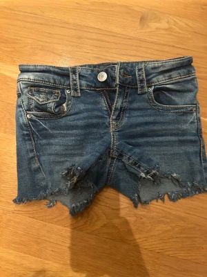 Hemma klippta Blå jeansshorts med slitna detaljer - Säljer ett par blå jeansshorts med råa, fransiga kanter och slitna detaljer framtill. Klassisk femficksmodell med knapp och dragkedja. Perfekta för en avslappnad och trendig look. Materialet är denim i bomull.