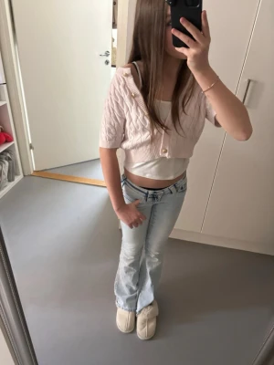 Ljusrosa kort kofta från Gina Tricot - Supersöt ljusrosa kort kofta från Gina Tricot med kabelstickat mönster och dekorativa knappar framtill. Perfekt att slänga över ett linne för en trendig lager-på-lager-look. Koftan har korta ärmar och mjukt stickat material.