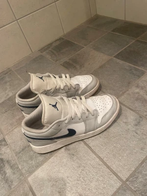 Nike Air Jordan 1 low  - Snygga vita sneakers från Nike Air Jordan 1 Low med marinblå detaljer och swoosh. Skorna har klassisk låg siluett, perforerad tåbox och vadderad plös med blå Jumpman-logga. Tillverkade i läder och textil för en clean och sportig look.