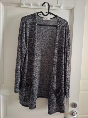 Melerad grå lång kofta med fickor - Säljer en lång, öppen kofta i melerad grå färg. Koftan har två praktiska fickor framtill. Perfekt att slänga över en t-shirt eller topp för en avslappnad look.
