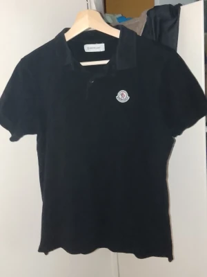 Moncler Svart pikétröja - Klassisk svart pikétröja från Moncler med korta ärmar, Moncler-logga på bröstet. Tröjan har krage och knappar vid halsen. Storlek S, kan passa M ifall man är lite smalare. T.ex denna tröjan passat mig länge å ja är 184cm och slim kroppsbyggnad.