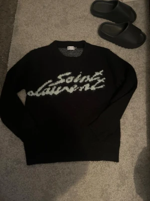 Svart stickad Saint Laurent tröja - Stickad svart tröja från Saint Laurent med vit fuzzy logga över bröstet. Med kvitto köpt för ungefär 10000kr.