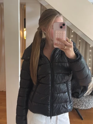 Svart pufferjacka med huva - Säljer en svart pufferjacka med dragkedja och avtagbar huva. Jackan har två fickor med dragkedja och quiltad design som ger en riktigt snygg och puffig look. Perfekt för kalla dagar och riktigt skön att bära. Passar till det mesta och är enkel att styla. 