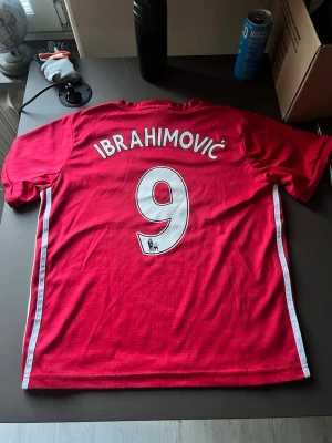 Manchester United Ibrahimović tröja - Röd Manchester United matchtröja med Ibrahimović och nummer 9 på ryggen. Tröjan har klubbmärke och Chevrolet-logga på bröstet samt vita detaljer längs sidorna. Perfekt för fotbollsfans som vill visa sitt stöd för laget.