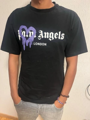 Svart Palm Angels t-shirt med tryck - Säljer en svart t-shirt från Palm Angels med stort vitt tryck på bröstet och ett lila hjärta med drip-effekt. T-shirten har klassisk passform och korta ärmar. Perfekt för dig som gillar streetwear och vill sticka ut med en unik detalj.