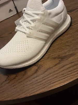 Vita Adidas Ultraboost sneakers - Säljer ett par helvita Adidas Ultraboost sneakers med stickad ovandel och klassiska tre ränder på sidan. Skorna har en bekväm Boost-sula och ventilerande meshmaterial. Perfekta för dig som gillar stilrena och sportiga sneakers.