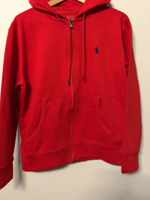 Röd zip hoodie från Polo Ralph Lauren - Röd hoodie med dragkedja från Polo Ralph Lauren. Klassisk modell med huva, dragsnören och kängurufickor framtill. Liten mörkblå logga broderad på bröstet. Mjuk bomullskänsla och ribbade muddar vid ärmar och nederkant.