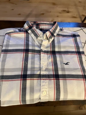 Rutig skjorta från Hollister - Säljer en klassisk rutig skjorta från Hollister i vitt, blått och rött. Skjortan har knappar framtill, button-down krage och det ikoniska Hollister-logotypen broderad på bröstet. Perfekt för en avslappnad men ändå stilren look.