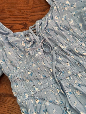 Blå blommig kortklänning med snörning - Söt ljusblå kortklänning med små vita och blå blommor. Klänningen har snörning vid bröstet, smock i midjan och volangdetaljer. Perfekt för sommarens varma dagar och har en luftig, feminin känsla.
