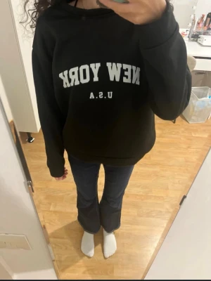 Svart sweatshirt med New York-tryck - Säljer en svart sweatshirt med vit text 'NEW YORK U.S.A.' på bröstet. Hoodien har en avslappnad passform och långa ärmar. Perfekt för dig som gillar streetwear och vill ha en enkel men snygg look.