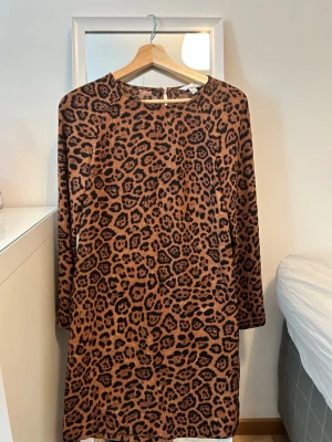 Leopardmönstrad långärmad klänning - Cool kortklänning med leopardmönster i bruna och svarta toner. Klänningen har rund halsringning och långa ärmar. Perfekt för dig som vill sticka ut med ett djurmönster och en avslappnad men trendig look.