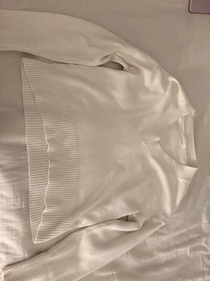 Vit stickad tröja från Gina Tricot - Säljer en stilren vit stickad tröja från Gina Tricot. Tröjan har v-ringning, långa ärmar och breda ribbade muddar vid ärmslut och nederkant. Perfekt för lager på lager och enkel att matcha med olika outfits.