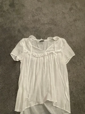 Vit luftig blus med volangdetaljer - Söt vit blus i tunn och luftig kvalitet med korta ärmar och volangdetaljer vid halsringningen. Blusen har knytband framtill och en avslappnad passform som ger en romantisk vibe. Perfekt till jeans eller kjol för en fräsch look.