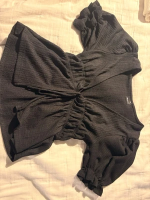 Svart blus med puffärm från Gina Tricot - Trendig svart blus från Gina Tricot i storlek XXS. Blusen har korta puffärmar, markerad midja med resår och en lätt rynkad struktur i tyget. Djup v-ringning och flowy passform ger en snygg siluett. Perfekt till jeans eller kjol.