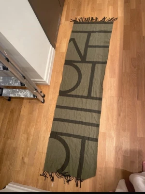 Olivgrön halsduk i ull från Totême - En ullhalsduk i olika gröna nyanser från Toteme. Superfin och  sparsamt använd!   Material: 100% ull  Mått: 173 x 48 cm  Skick: jättefint, några enstaka fransar har gått upp lite (se bild), i övrigt inget att anmärka.