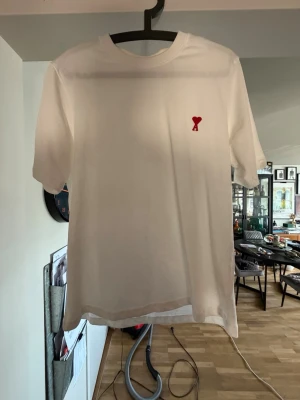 Vit t-shirt från Ami Alexandre Mattiussi - Vit t-shirt från Ami Alexandre Mattiussi med rakt snitt och korta ärmar. På bröstet finns ett litet rött hjärta med bokstaven A. Tillverkad i mjuk bomull som känns skön mot huden. Perfekt till jeans eller shorts för en clean look. Obs kvitto finns ej då den e köpt utomlands