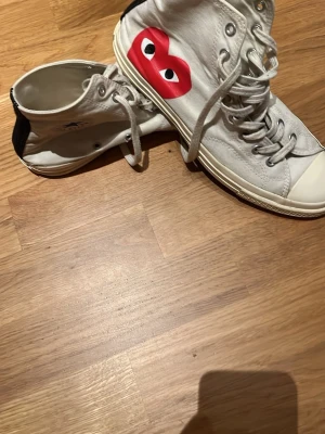 Vita höga sneakers med röd hjärtlogga - Säljer ett par vita höga sneakers med den ikoniska röda hjärtloggan med ögon på sidan. Skorna har vita snören, gummisula och svart detalj på hälen. Klassisk canvasmodell som sticker ut med sin coola design.