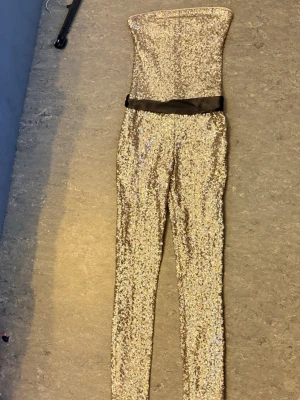 Guldig paljettjumpsuit med svart band - Snygg axelbandslös jumpsuit i guld med glittrande paljetter över hela plagget. Tight passform och långt ben, dekorerad med ett svart band i midjan. Perfekt för dig som vill sticka ut och glänsa. Materialet är stretchigt och följsamt.