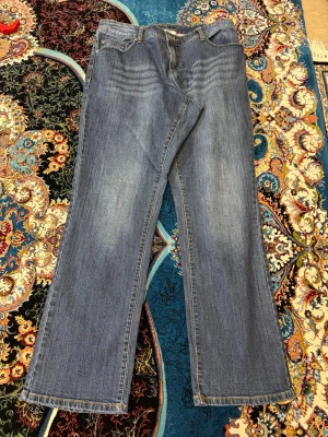 Blå bootcut jeans med slitningar - Säljer ett par klassiska blå bootcut jeans med snygga slitningar och ljusare partier framtill. Jeansen har fem fickor och normal midja. Perfekta för en avslappnad och trendig look. Materialet är denim i bomull.