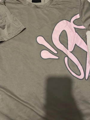 Synaworld t shirt centralcee  - Säljer en beige t-shirt med ett stort rosa grafiskt tryck på framsidan. T-shirten har korta ärmar och rund hals. Materialet känns mjukt och skönt, troligtvis bomull. Perfekt för en chill och avslappnad stil.