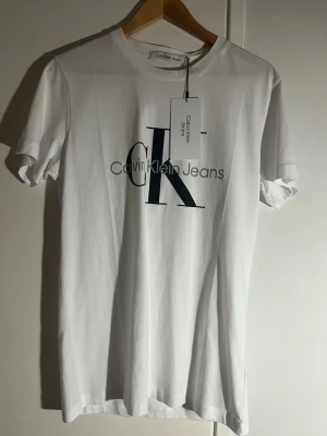 Vit Calvin Klein Jeans t-shirt - Snygg vit t-shirt från Calvin Klein Jeans med stor CK-logga och text på bröstet. Klassisk passform med rund hals och korta ärmar. Perfekt till jeans eller shorts för en clean och stilren look.