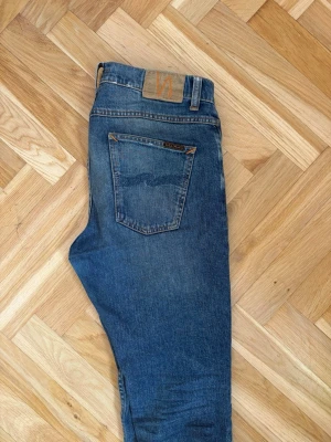Blå jeans från Nudie Jeans - Säljer ett par klassiska blå jeans från Nudie Jeans med raka ben och snygga detaljer på bakfickorna. Jeansen har en mellanblå tvätt och är tillverkade i denim med en skön passform. Perfekta för en avslappnad stil.