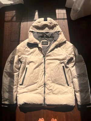Beige vindjacka från Sail Racing - Snygg beige vindjacka från Sail Racing med svart dragkedja och detaljer. Jackan har två framfickor med dragkedja, hög krage och huva. Materialet är slitstarkt och vattenavvisande, perfekt för blåsiga dagar. Logga på bröstet och ärmen för en sportig look.