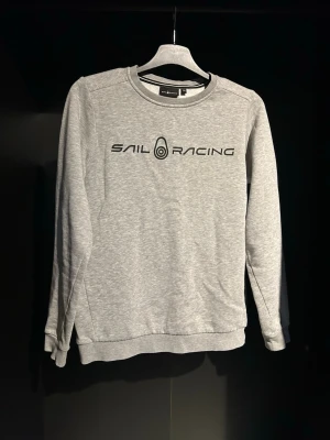 Grå sweatshirt från Sail Racing - Snygg grå sweatshirt från Sail Racing med svart logotyptryck på bröstet och en liten logga i nacken. Tröjan har rund halsringning, långa ärmar och ribbade muddar. Perfekt för en avslappnad och sportig stil. Strlk 150