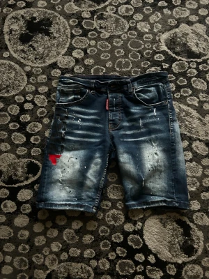Blå jeansshorts med tryck - Säljer ett par blå jeansshorts med coolt sliten look, vita färgstänk och svart tryck på vänster ben. Det finns även en röd detalj på samma ben. Shortsen har fem fickor och knappgylf. Perfekta för sommaren!