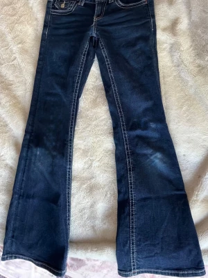 Mörkblå bootcut jeans - Snygga mörkblå bootcut jeans med vita kontrastsömmar och klassiska fem fickor. Jeansen har utsvängda ben och dekorativa ficklock med knapp bak. Perfekta för dig som gillar retrostil och vill ha ett par jeans som sticker ut.