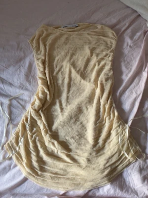 Ljusgul kort blus från ZARA - Säljer en ljusgul kort blus från ZARA med tunna horisontella ränder och snörning i sidorna. Klänningen har en enkel, rak form och är tillverkad i ett lätt, tunt material som känns perfekt för varmare dagar.