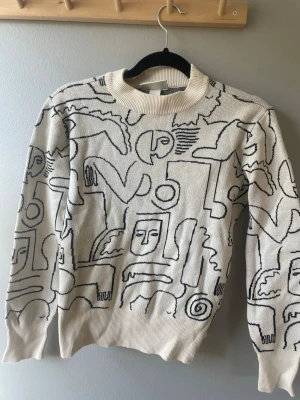 Beige stickad tröja med grafiskt mönster - Snygg beige stickad tröja från Monki med svart grafiskt mönster av ansikten och former. Tröjan har rund halsringning och ribbade muddar vid ärmslut och nederkant. Perfekt för dig som gillar unik och konstnärlig stil.