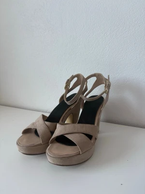 Beige platåsandaler med klack - Snygga beige sandaler med hög klack och platåsula. Skorna har korsade remmar framtill och justerbart spänne vid ankeln i guld. Tillverkade i mockaimitation med svart innersula. Perfekta för dig som vill ha både höjd och stil.