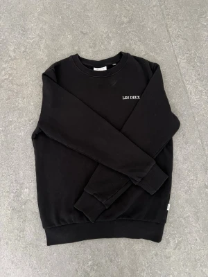 Svart sweatshirt från Les Deux - Säljer en svart sweatshirt från Les Deux med rund halsringning och diskret vit logga på bröstet. Tröjan har ribbade muddar vid ärmslut och nederkant. Perfekt för en clean och stilren look.