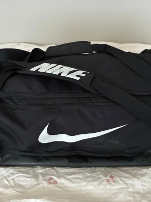 Svart sportbag från Nike - Snygg svart sportbag från Nike med stort vitt logotryck på sidan och axelremmen. Väskan har dragkedja, justerbar axelrem med vadderad del och flera fack för smart förvaring. Perfekt för gymmet eller resan.
