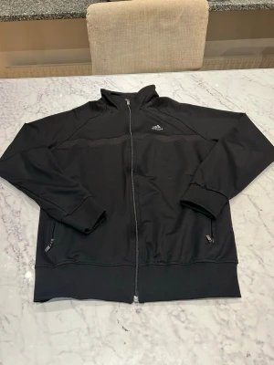 Svart ziptröja från Adidas - Svart ziptröja från Adidas med hög krage och dragkedja framtill. Tröjan har två fickor med dragkedja, ribbade muddar och CLIMALITE-material som andas. Adidas-logga på bröstet och diskret CLIMALITE-tryck bak. Perfekt till sport eller chill.