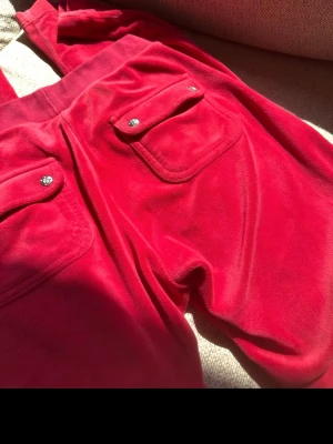 Röda  Juicy Couture - Säljer ett par klassiska röda mjukisbyxor från Juicy Couture. Byxorna har två fickor bak med lock och blingiga knappar. Perfekta för en chill och trendig look. Elastisk midja för skön passform. Byxorna har ett litet hål bredvid ena fickan som man kan se på bild 3. Dom har inte heller nått snöre💕
