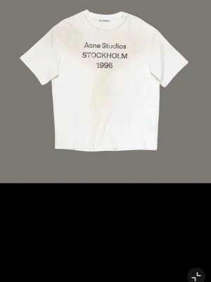 Vit Acne Studios t-shirt med tryck - Snygg vit t-shirt från Acne Studios med svart texttryck på bröstet där det står 'Aone Studios STOCKHOLM 1996'.observera att det står aone istället för acne. Klassisk rak passform och rund halsringning. Perfekt till jeans eller shorts för en clean och trendig look.Storlek M passar S