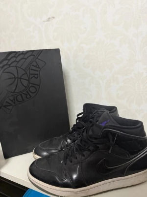 Svarta Air Jordan 1 Mid sneakers - Säljer ett par svarta Air Jordan 1 Mid sneakers med lila Jumpman-detalj på plösen. Skorna har klassisk siluett, svart läderöverdel och vit sula. Swoosh-logga på sidan och svart snörning. Box medföljer.