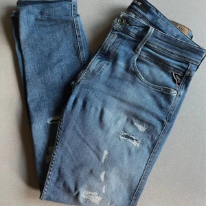 Replay Anbass blå jeans, storlek 32/30 - Snygga Replay Anbass jeans i klassisk blå tvätt med slitningar och lätt distressed detaljer. Modellen har fem fickor, normal passform och raka ben. Jeansen har ett hål i skrevet. Jeansen är tillverkade i mjukt denimtyg med stretch för extra komfort. Perfekt för dig som gillar en avslappnad och modern look.
