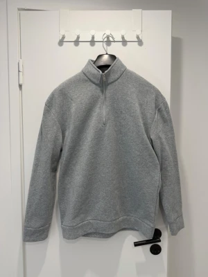 Grå halvzip hoodie - En stilren grå hoodie med halv dragkedja och hög krage. Tillverkad i mjukt sweatshirtmaterial som känns skönt mot huden. Långärmad modell utan synliga loggor eller tryck, perfekt för en clean och enkel look.
