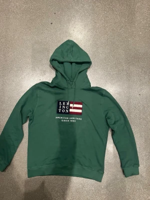 Grön hoodie från Lexington - Mörkgrön hoodie från Lexington med broderad amerikansk flagga och texten 'American Heritage Since 1993' på bröstet. Hoodien är i storlek 170 men skulle säga att den sitter som XS. Den är i princip alldrig använd så är i helt nyskick!                                     Priset är går absolut att diskutera!