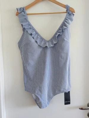 Randig baddräkt med volangdetaljer - Snygg baddräkt med smala blå och vita ränder och volang längs ringningen och axelbanden. Djup V-ringning både fram och bak ger en trendig look. Tillverkad i ett stretchigt material som sitter bekvämt på kroppen. Perfekt för strand eller poolhäng.