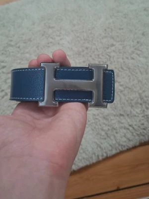 Blått skärp med stor H-spänne - Snyggt blått skärp med bred rem och ett stort metallspänne i form av bokstaven H. Skärpet har vita kontrastsömmar längs kanterna och känns riktigt stilrent. Perfekt accessoar för att lyfta din outfit.