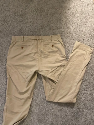Beige chinos från Polo Ralph Lauren - Snygga beige chinos från Polo Ralph Lauren med klassisk rak passform. Byxorna har två bakfickor med knappar, bälteshällor och en diskret Polo-logga ovanför ena bakfickan. Tillverkade i ett mjukt bomullsmaterial som känns bekvämt hela dagen. Storlek = 30/34