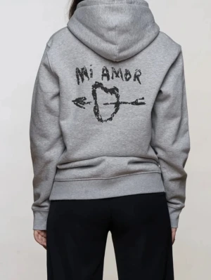 Grå hoodie med tryck 'Mi Amor' - Säljer en grå hoodie med huva och ett svart tryck på ryggen där det står 'Mi Amor' och ett hjärta genomborrat av en pil. Hoodien har en avslappnad passform och är tillverkad i mjukt sweatshirtmaterial. Perfekt för dig som gillar streetwear och unika prints.