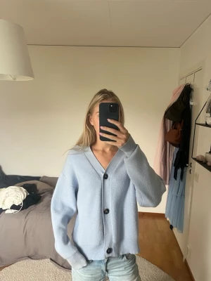 Ljusblå oversized kofta - Mysig ljusblå kofta med stora svarta knappar och v-ringad hals. Koftan har en oversized passform, långa ärmar och ribbade muddar vid ärmslut och nederkant. Perfekt för lager på lager och en chill look.