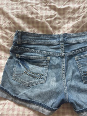 Blå jeansshorts med nitar - Säljer ett par klassiska blå jeansshorts med uppvikta benslut och snygga nitar på bakfickorna. Shortsen har normal passform, bälteshällor och knappstängning framtill. Perfekta för varma dagar och enkel att matcha med allt.